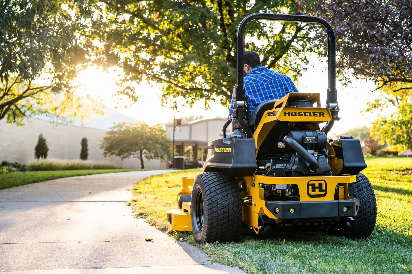 Person operating a Hustler zero-turn lawn mower in a park-like setting.