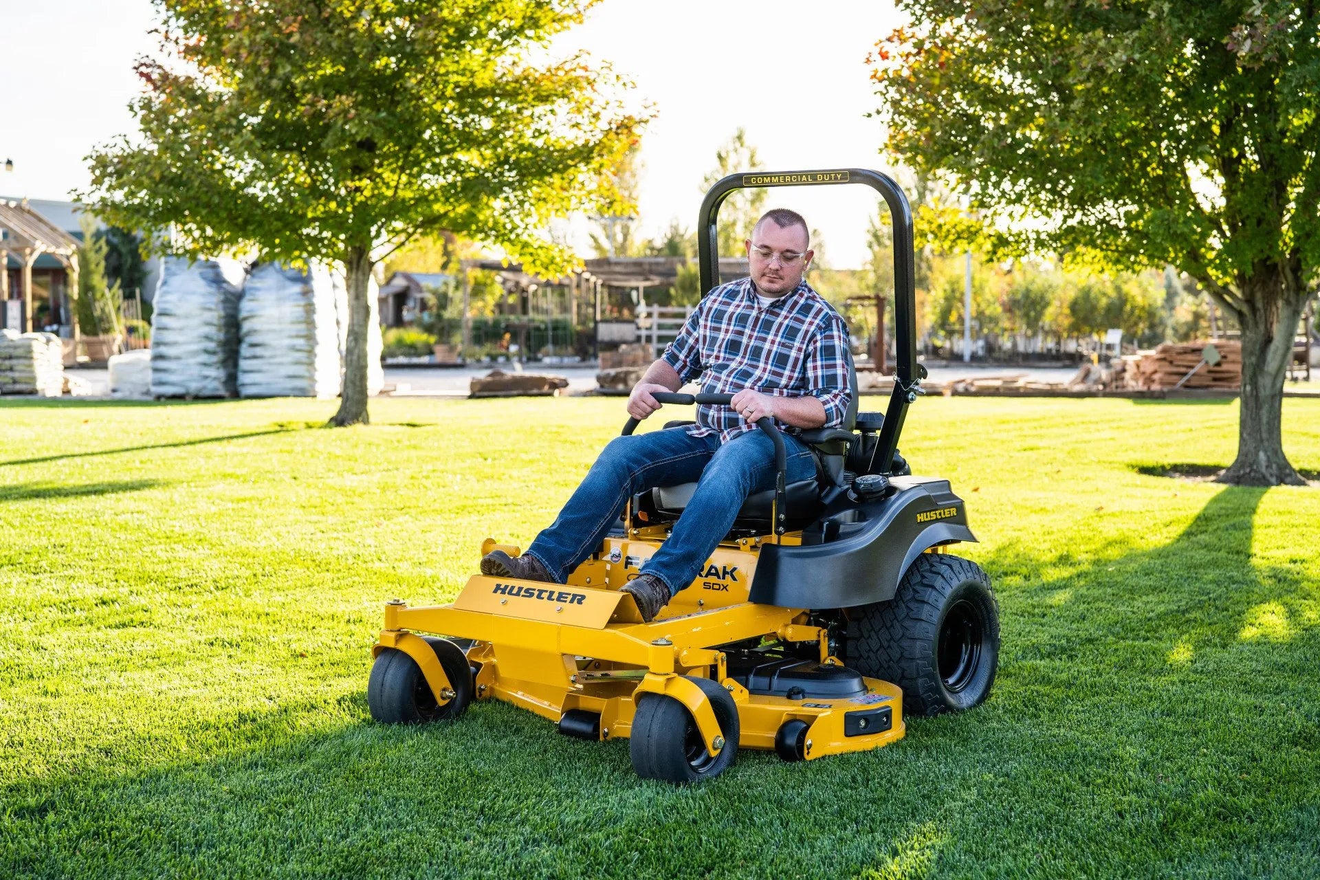 Man operating a yellow Hustler mower on a grassy area with trees and buildings in the background.