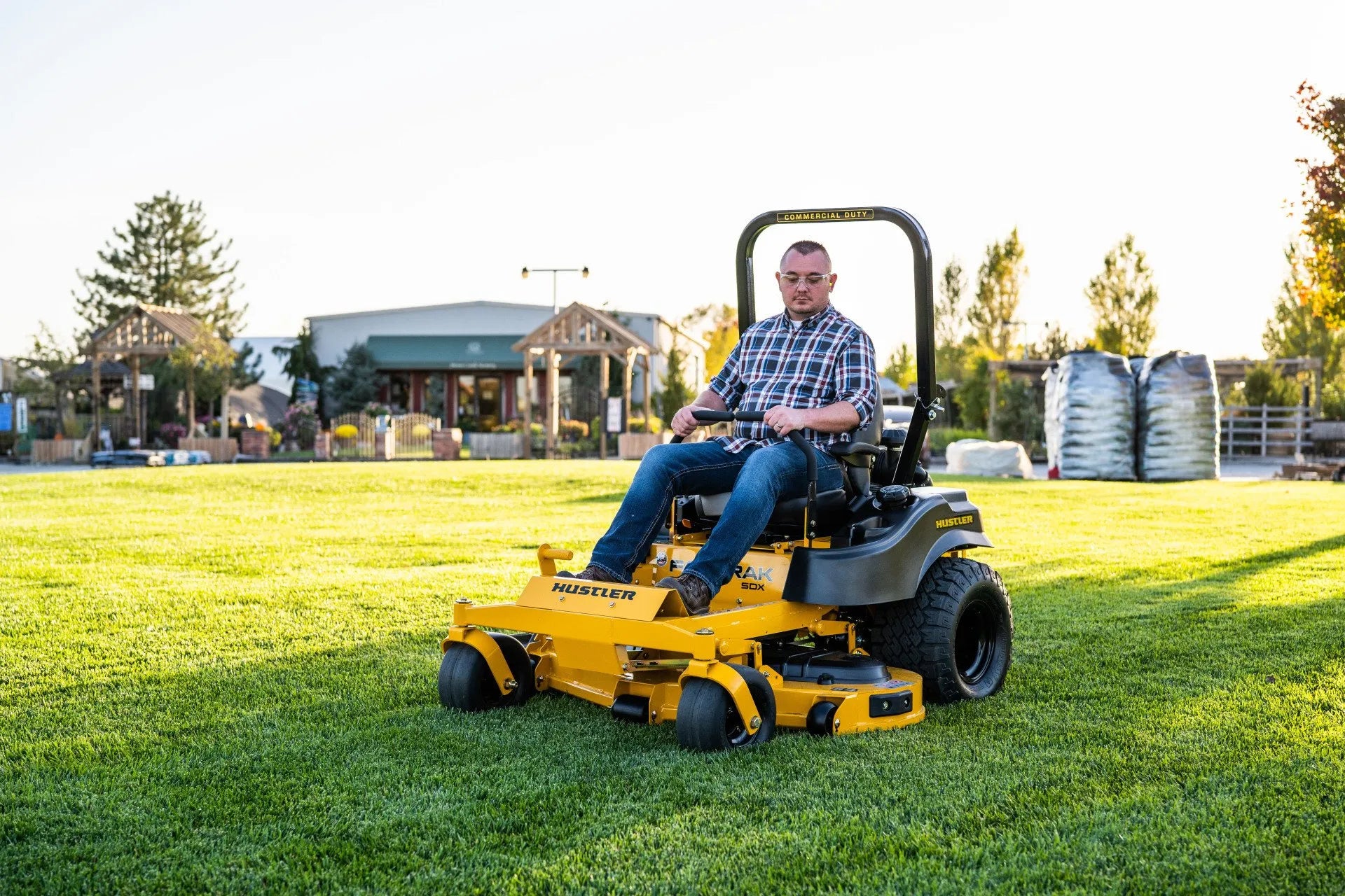 Man operating a yellow riding lawn mower on a grassy field with buildings and trees in the background.