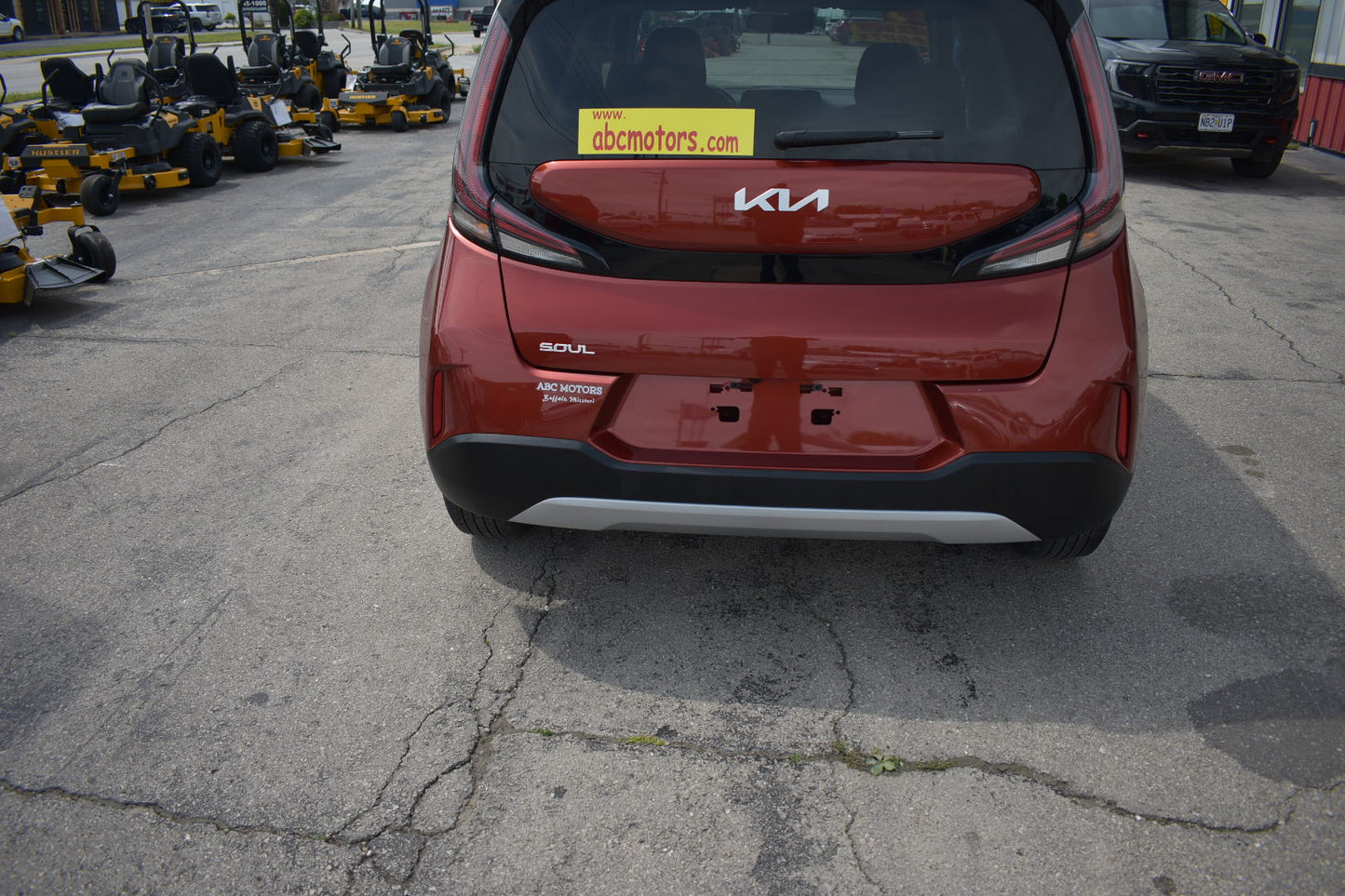 Red Kia Soul car parked on a cracked asphalt surface with lawn mowers in the background.