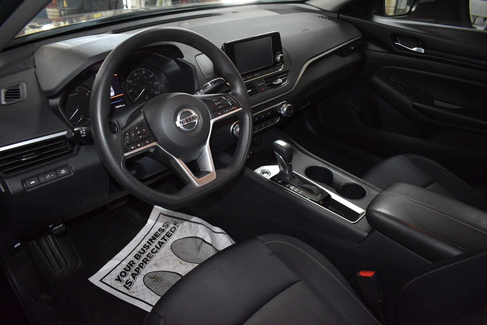 Car interior with steering wheel and dashboard, featuring a humorous doormat on the floor.