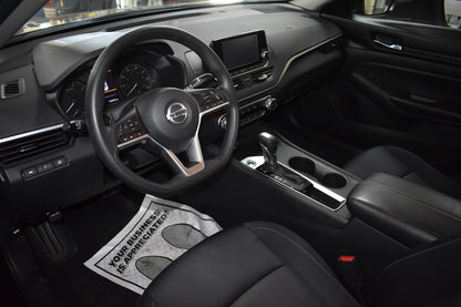 Car interior with steering wheel and dashboard, featuring a humorous doormat on the floor.