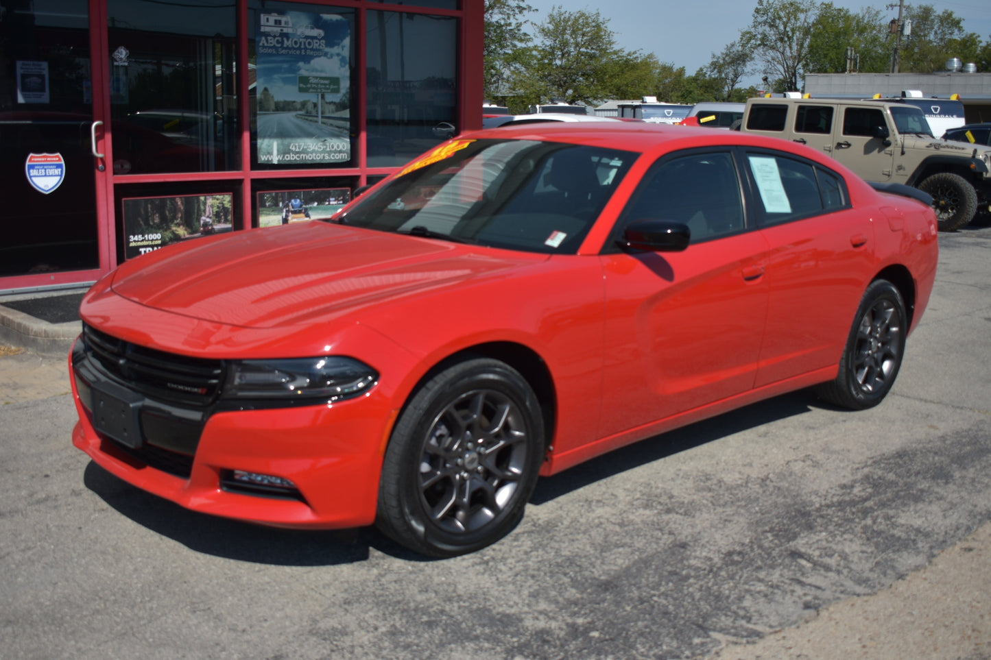 A bright red Dodge Charger sedan parked on a dealership lot in front of a red building, with other vehicles visible in the background.