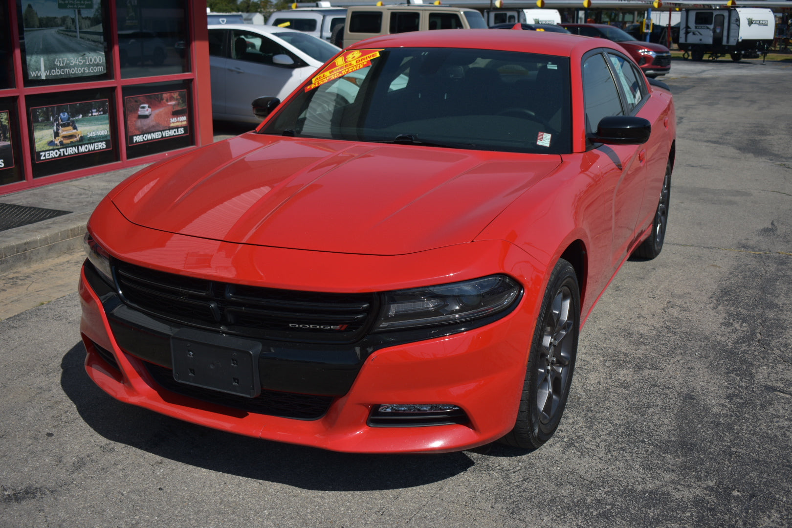 A bright red Dodge Charger sedan parked outdoors on a paved lot in front of a dealership building. The front grille and headlights are visible, along with a yellow price tag on the windshield.