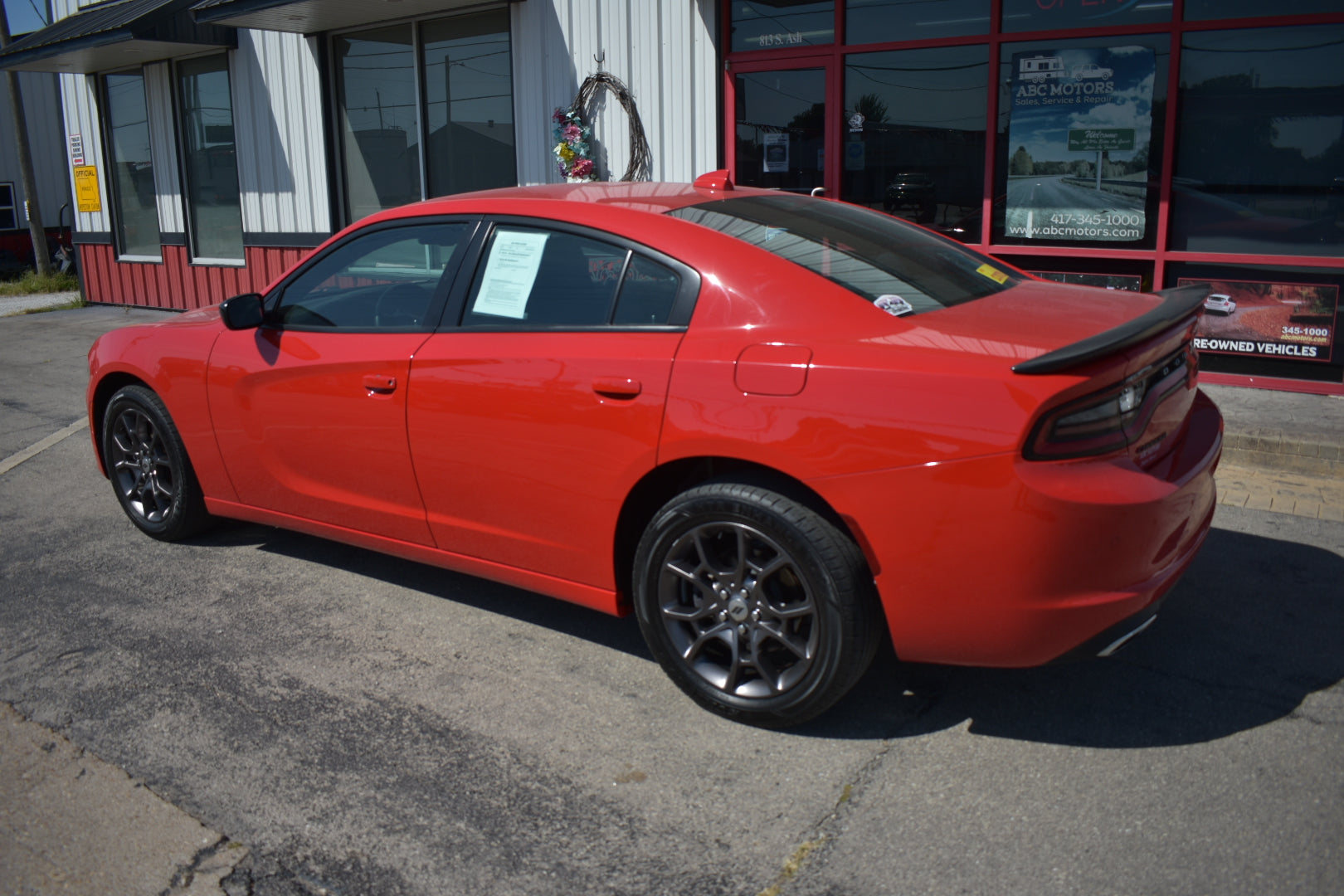A bright red Dodge Charger sedan parked outdoors in front of a dealership building with large windows and a red frame.