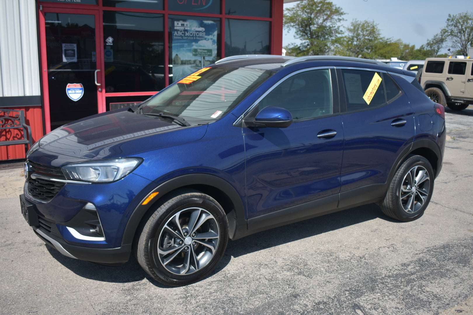 A dark blue buick encore parked on an outdoor dealership lot, with a red and white building, trees, and other vehicles visible in the background.