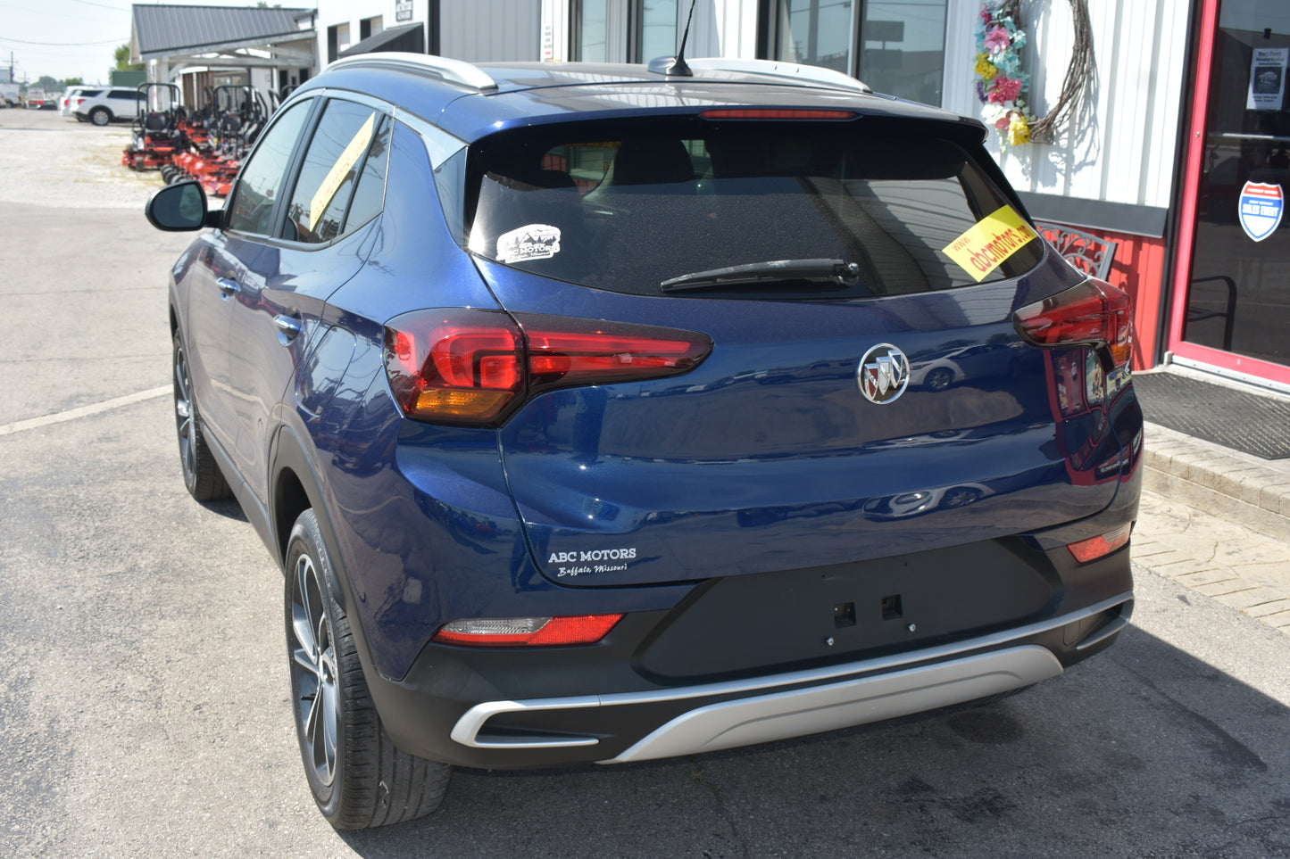 A blue Buick Encore SUV parked outdoors in front of a dealership building, shown from the rear three-quarter angle. The vehicle features a dark metallic blue paint job, a silver chrome trim strip on the rear bumper, and a visible Buick logo in the center.