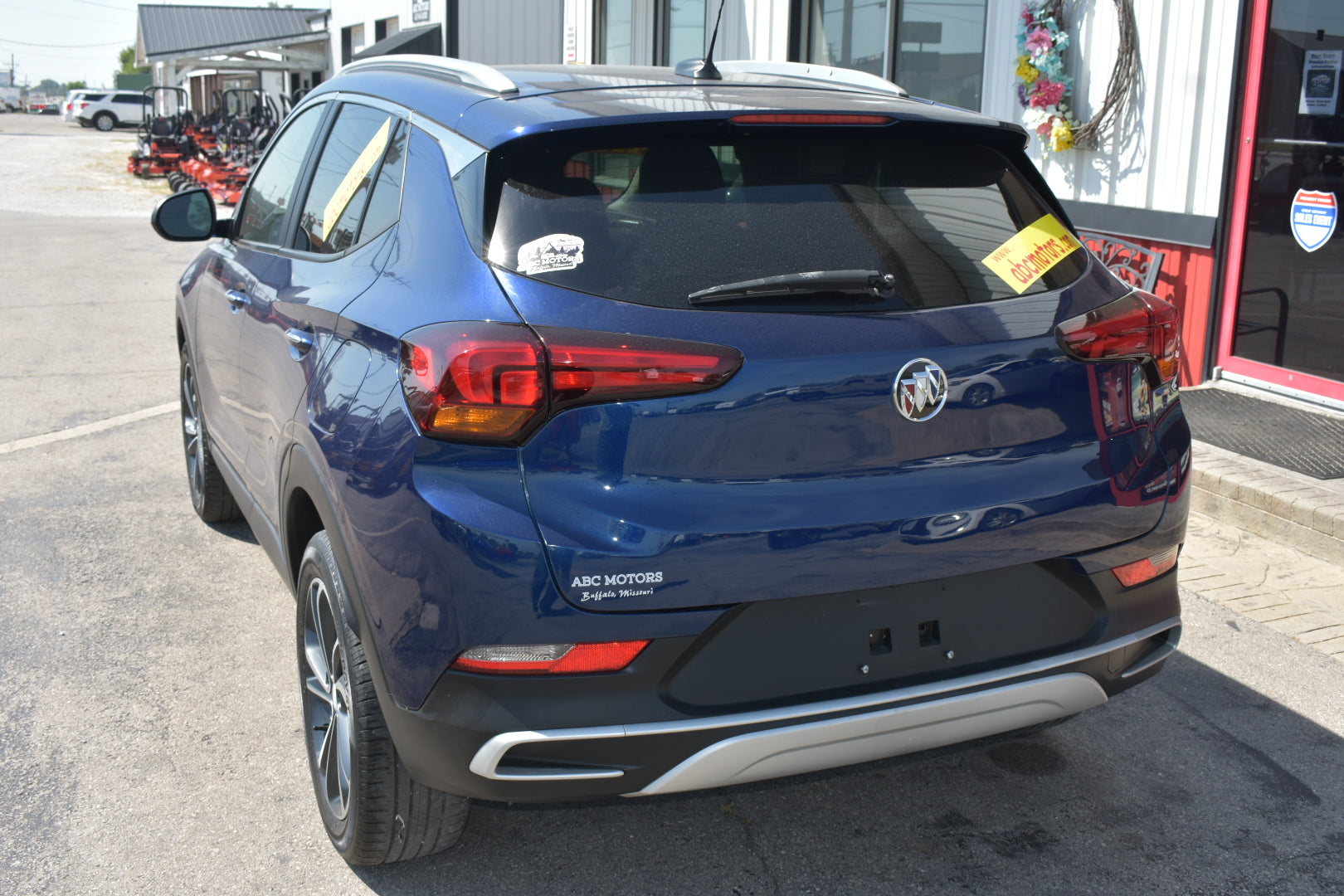 A blue Buick Encore SUV parked outdoors in front of a dealership building, shown from the rear three-quarter angle. The vehicle features a dark metallic blue paint job, a silver chrome trim strip on the rear bumper, and a visible Buick logo in the center.