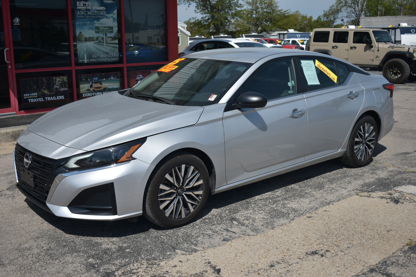 A silver Nissan sedan parked on a dealership lot, with a red building and other vehicles visible in the background. Yellow price stickers are attached to the windshield.