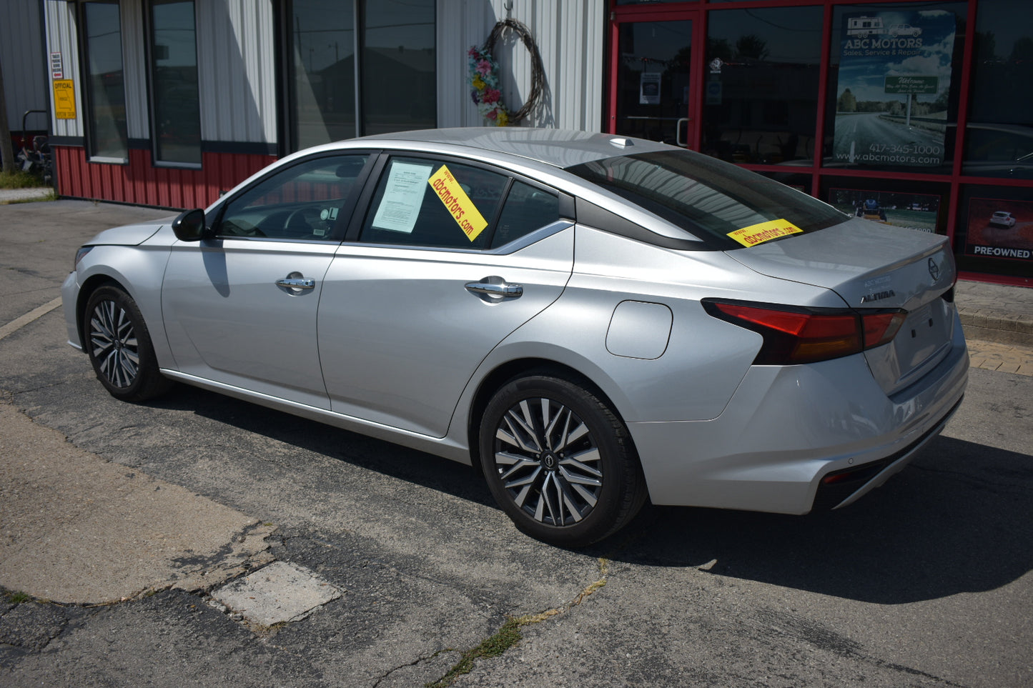 A silver Nissan Altima sedan parked outdoors in front of a dealership building, shown from a rear three-quarter angle with yellow sales stickers on the windows.