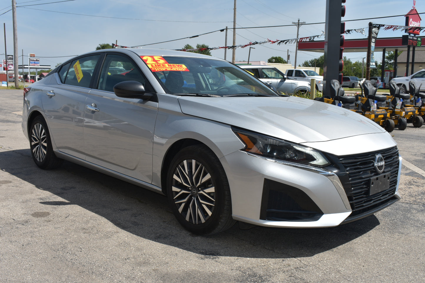 A silver Nissan sedan parked outdoors in a dealership lot, with a yellow price sticker on the windshield and other cars and equipment visible in the background.