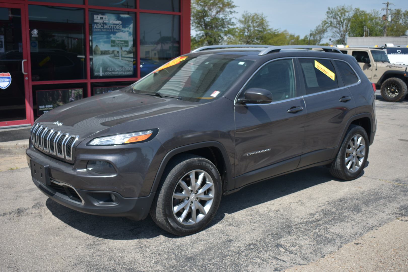 A dark gray Jeep Cherokee SUV parked on an asphalt lot in front of a red-framed dealership building, with yellow dealership stickers on the windshield.