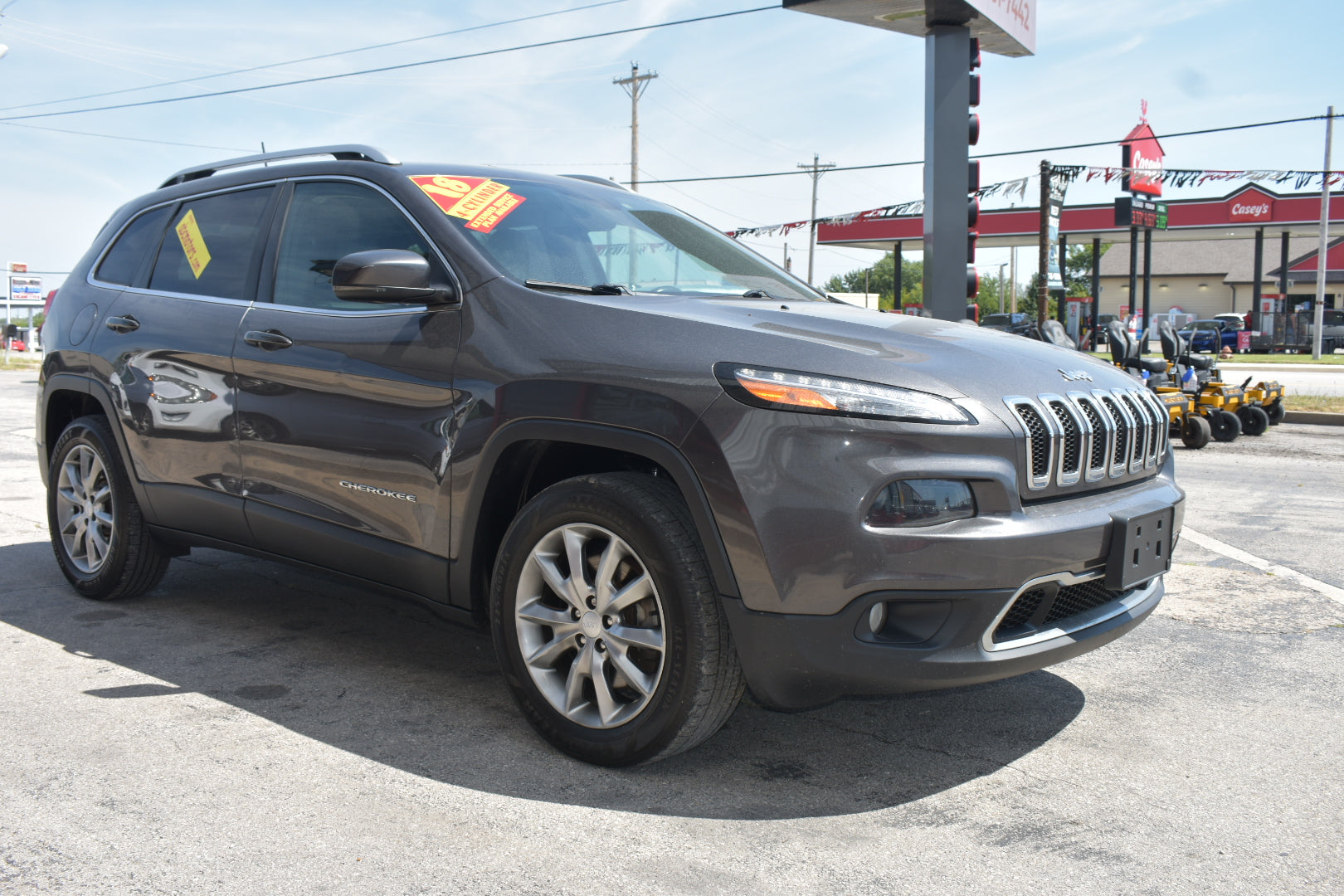 A dark gray 2018 Jeep Cherokee SUV parked on an asphalt lot at a dealership, with a red and yellow sales sticker on the windshield and a gas station in the background.