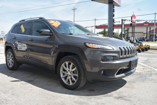 A dark gray 2018 Jeep Cherokee SUV parked on an asphalt lot at a dealership, with a red and yellow sales sticker on the windshield and a gas station in the background.
