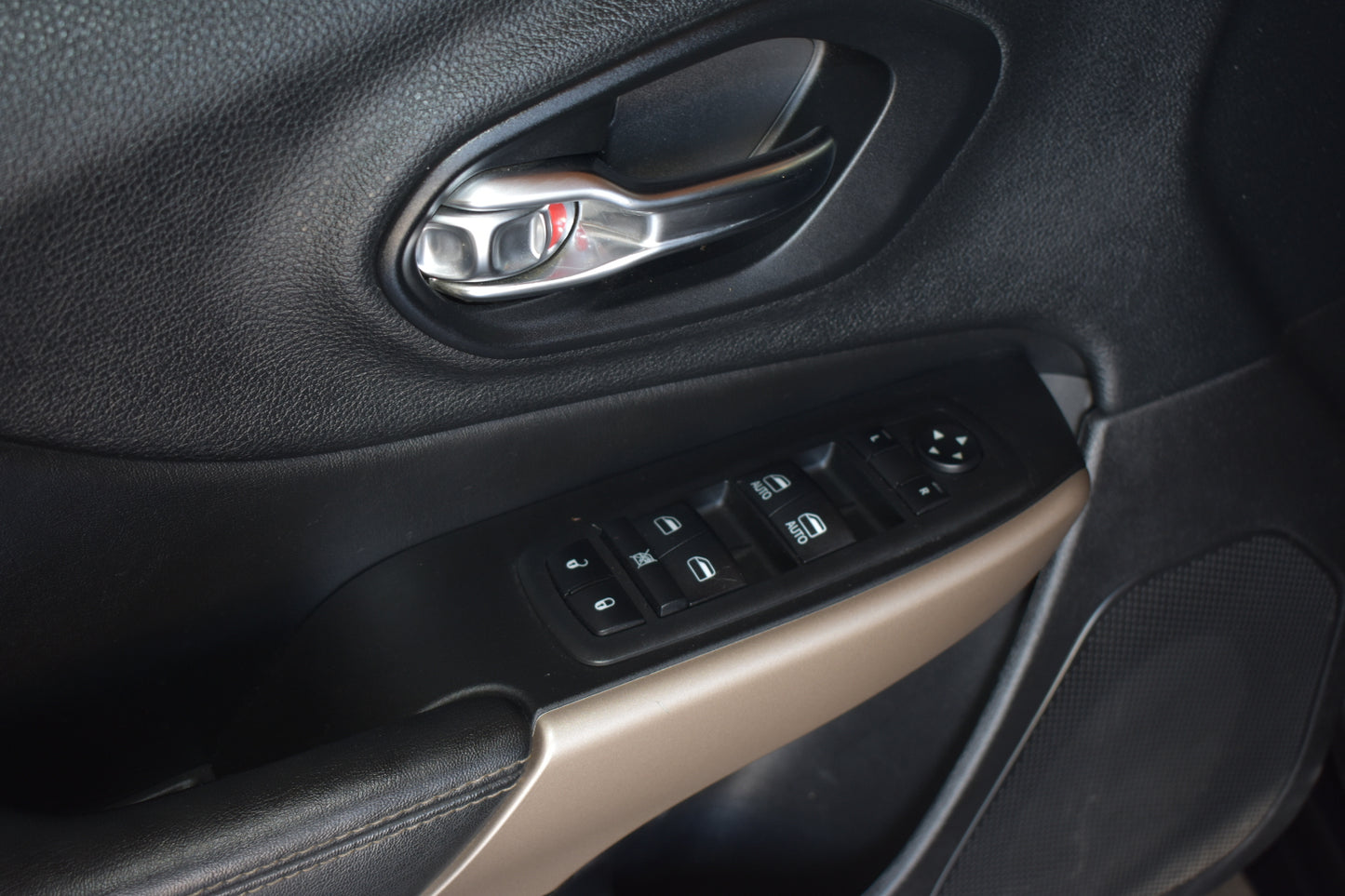 Close-up of a car door panel showing the power window and door lock controls, with a silver door handle above and a beige trim accent below.