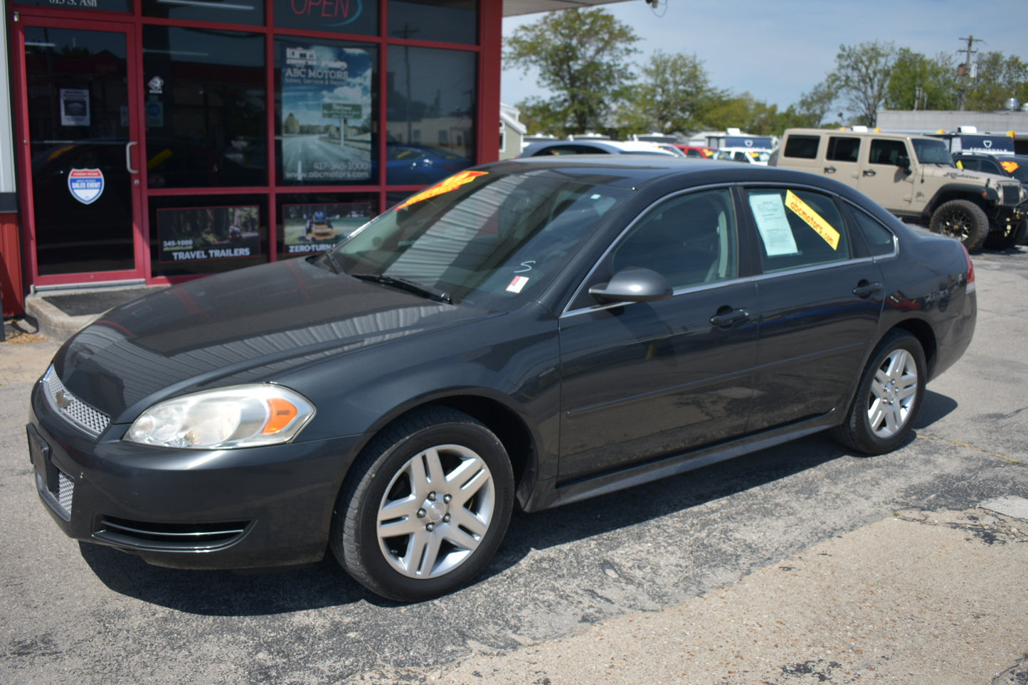 A dark gray 2008 Chevrolet Impala sedan parked on a dealership lot in front of a red building with glass windows. Yellow sales stickers are visible on the windshield and side window.