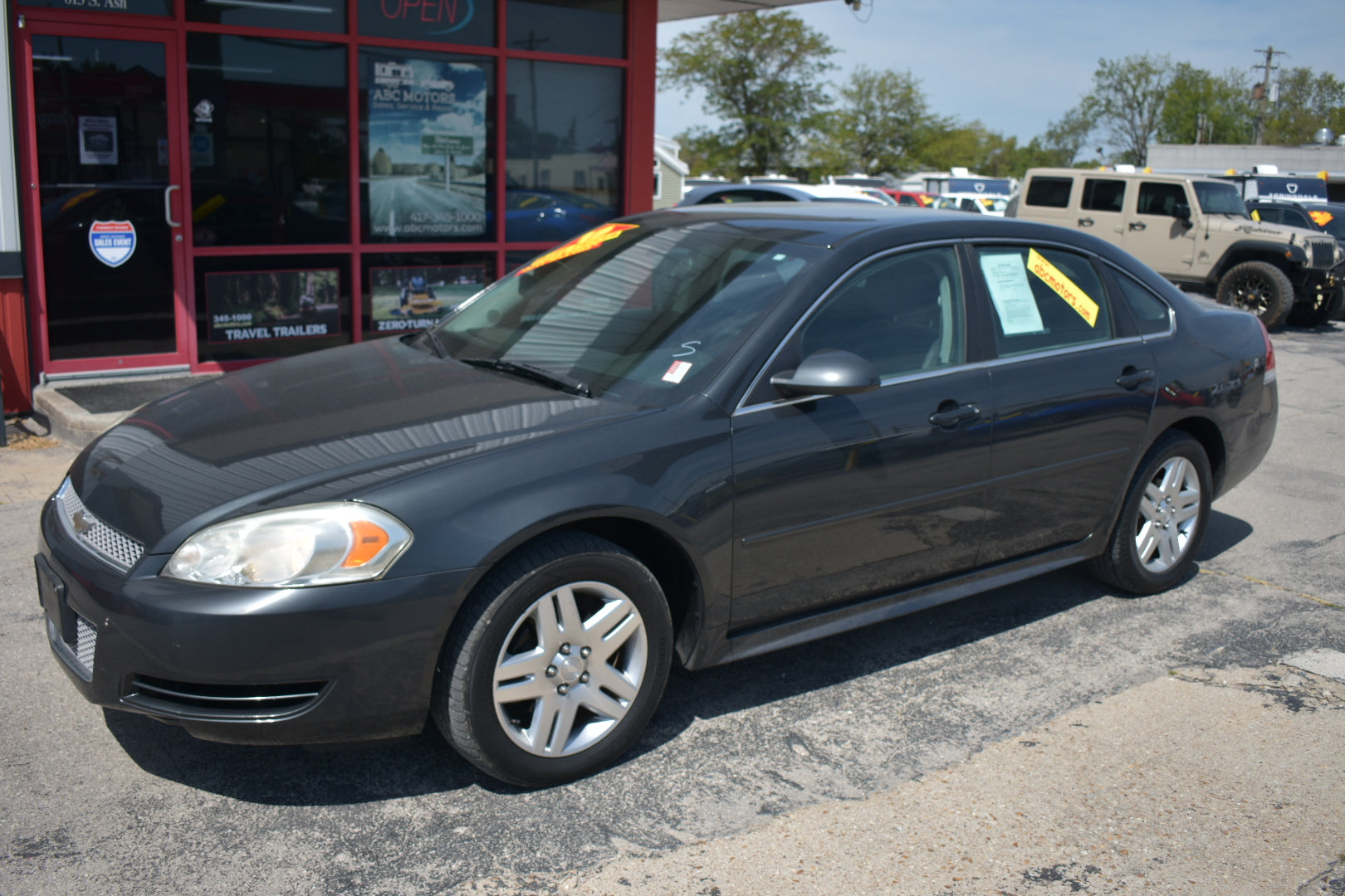 A dark gray 2008 Chevrolet Impala sedan parked on a dealership lot in front of a red building with glass windows. Yellow sales stickers are visible on the windshield and side window.