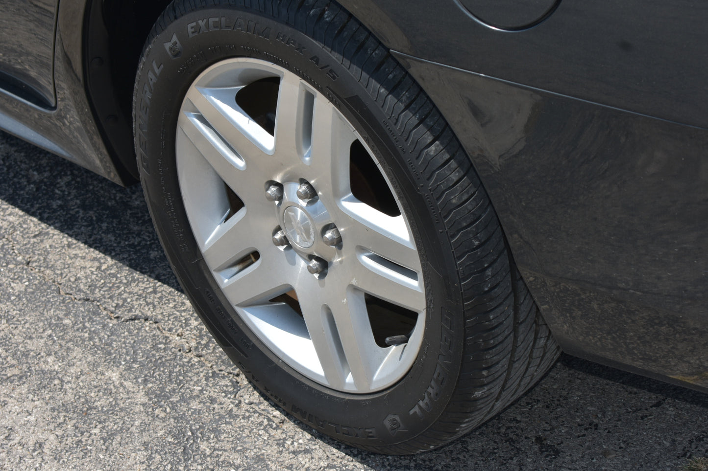 A close-up shot of a silver multi-spoke alloy wheel and black tire mounted on a dark gray car, parked on asphalt.