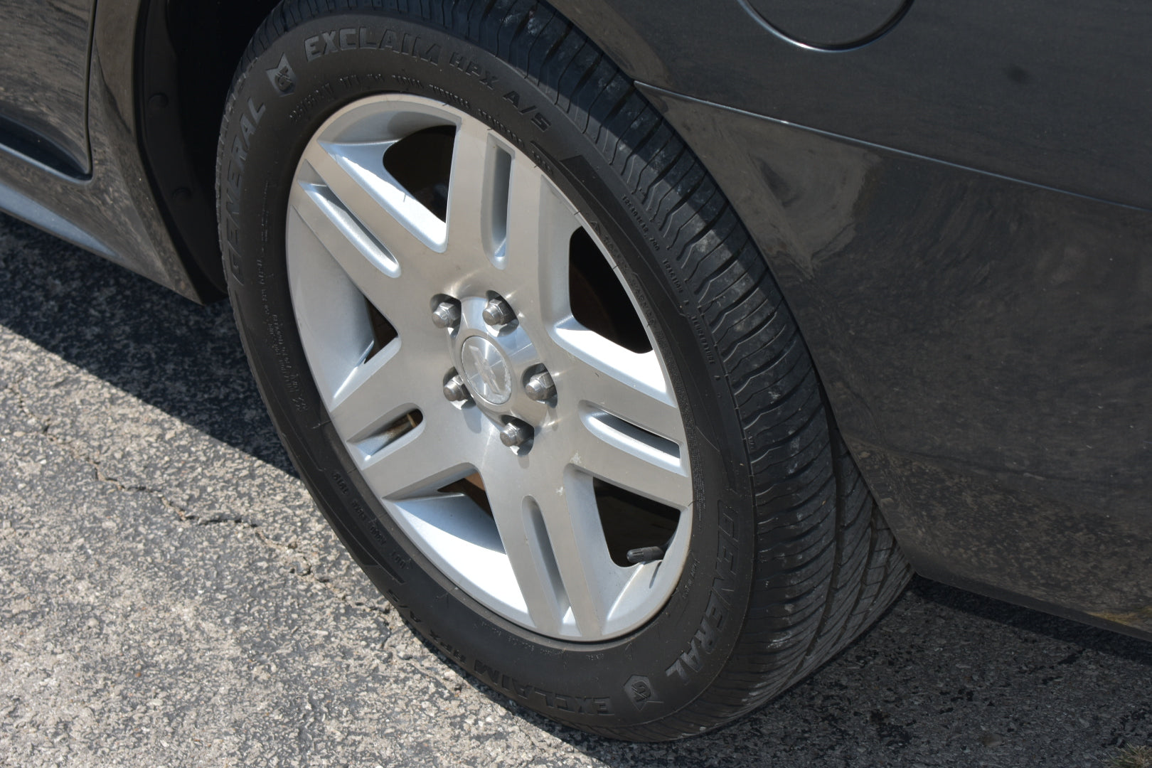 A close-up shot of a silver multi-spoke alloy wheel and black tire mounted on a dark gray car, parked on asphalt.