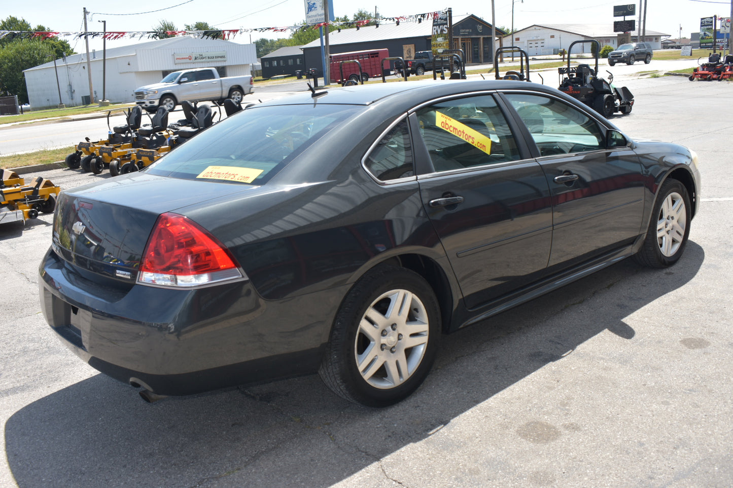 A black sedan parked outdoors in a dealership lot, photographed from the rear three-quarter angle. The car has silver multi-spoke wheels and yellow sales stickers on the rear and side windows.