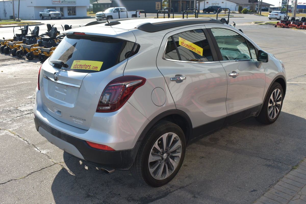 A silver Buick Encore SUV parked on an outdoor dealership lot, viewed from a rear three-quarter angle. The vehicle has a yellow 'For Sale' sign on the rear window and a matching sign on the side window.