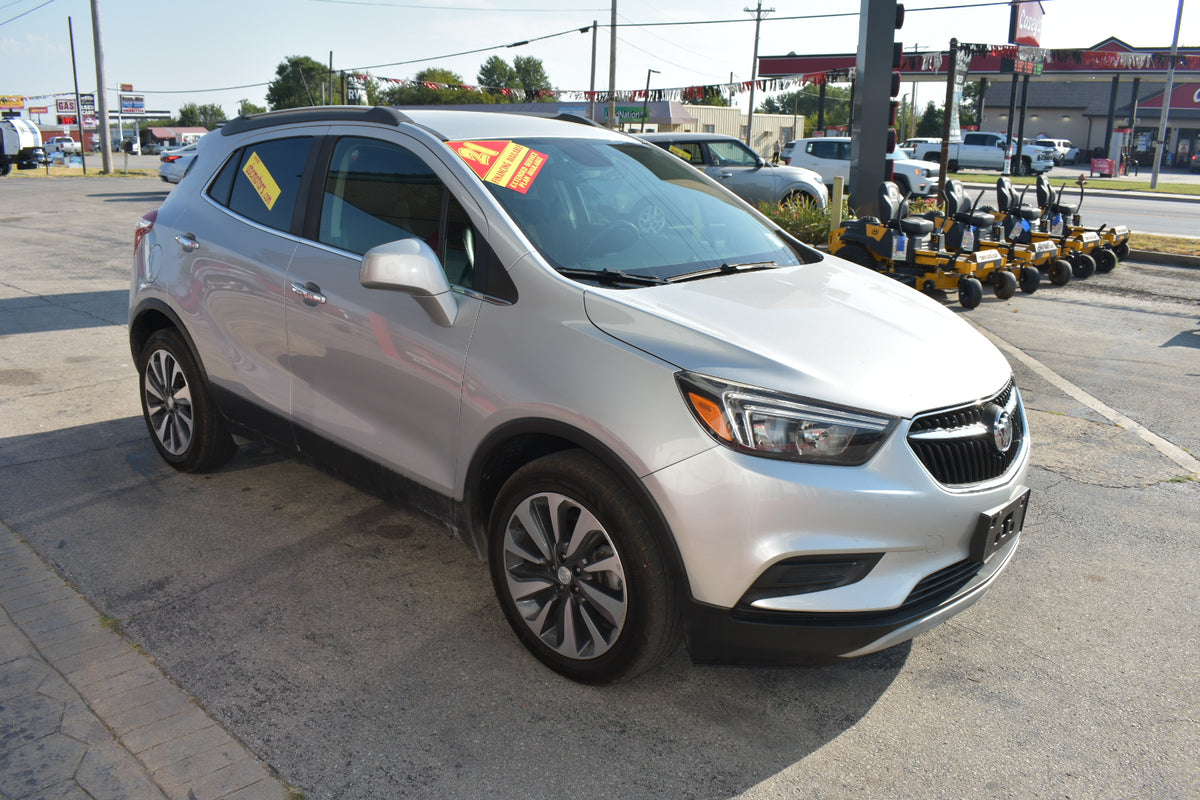 A silver Buick Encore SUV parked in an outdoor dealership lot, with a red and yellow 'For Sale' sticker on the windshield. The vehicle is photographed from a three-quarter front angle, showing its front grille, headlights, alloy wheels, and side profile.