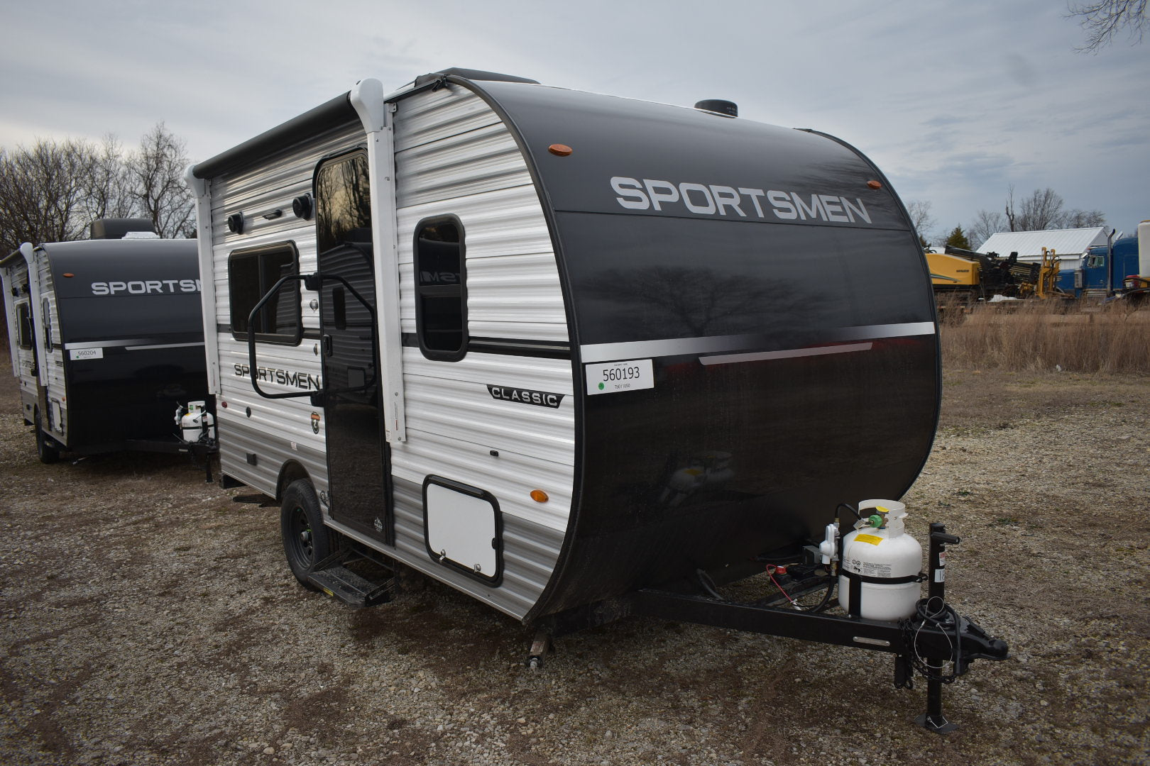 Two 'Sportsmen' travel trailers parked on a gravel lot with a cloudy sky.