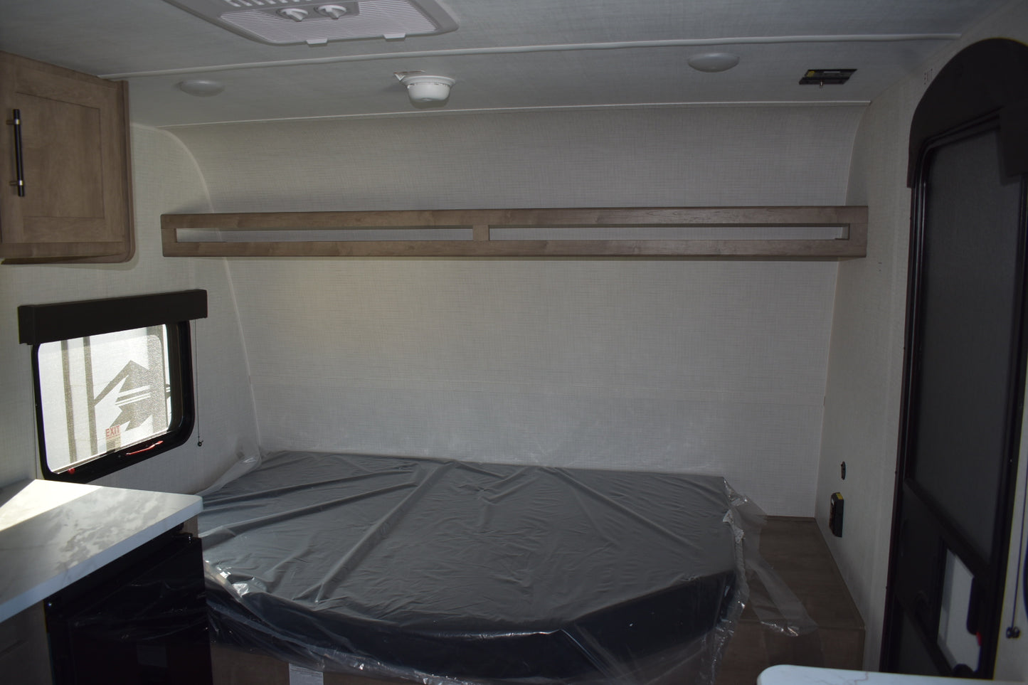 Compact interior of a vehicle with a bed covered in plastic wrap, wooden shelf, and small window.