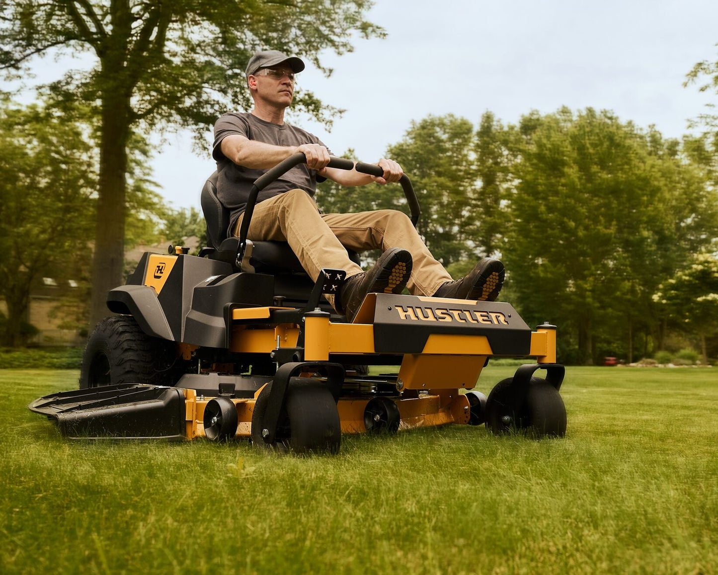 Man operating a Hustler lawn mower in a grassy area with trees in the background