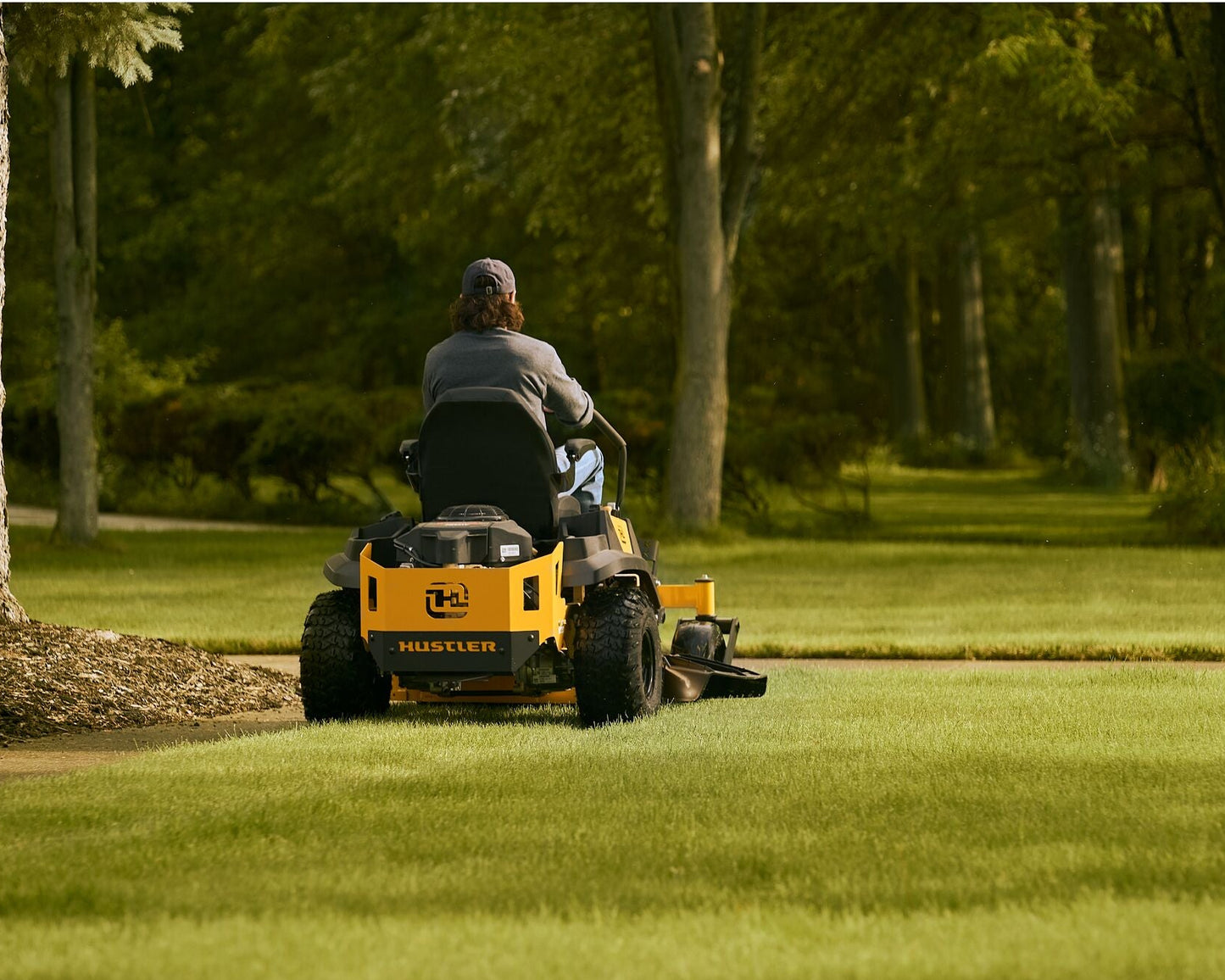 Person operating a yellow riding lawn mower in a park-like setting with trees and grass.
