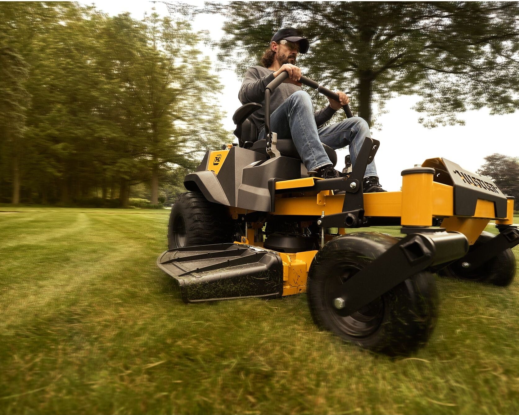 Person operating a yellow riding lawn mower on a grassy field with trees in the background