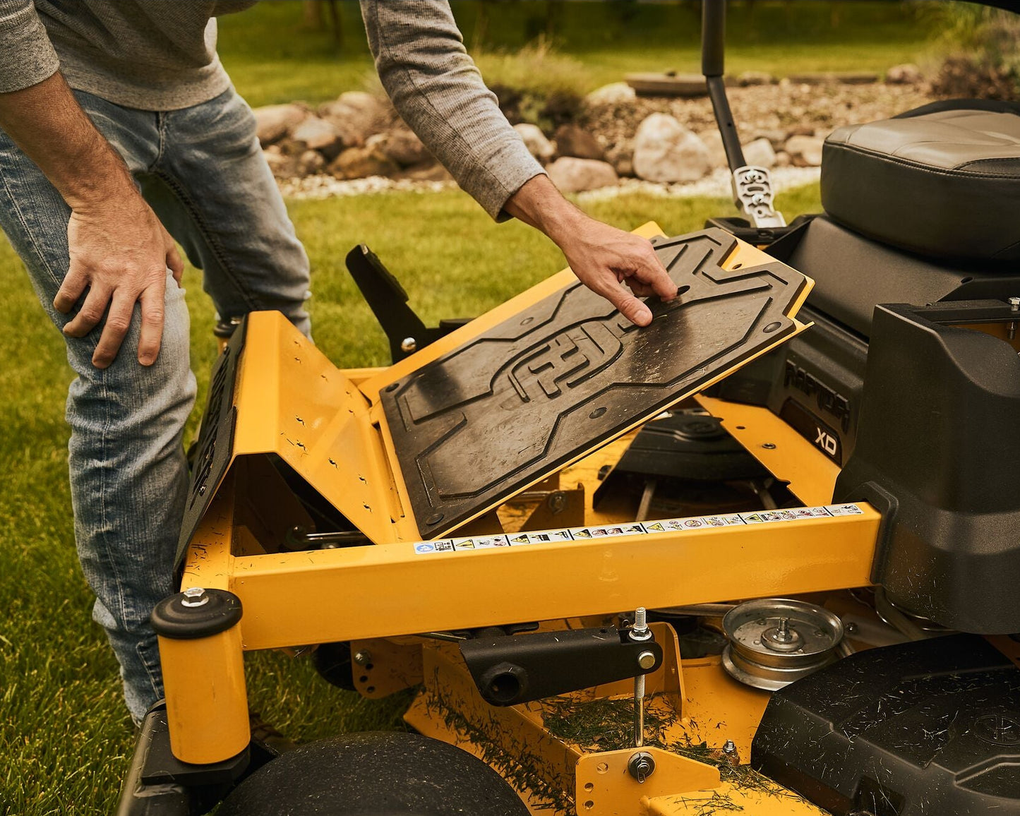 Person adjusting a lawn mower deck on a riding lawn mower with a grassy area in the background.