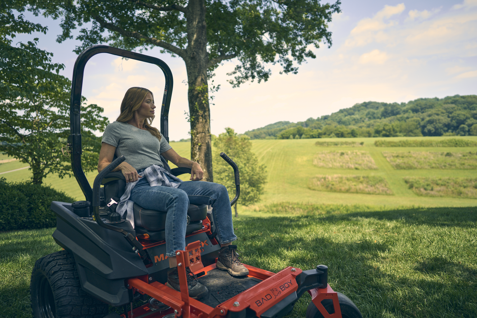 Woman sitting on a lawn mower in a grassy field with trees and mountains in the background